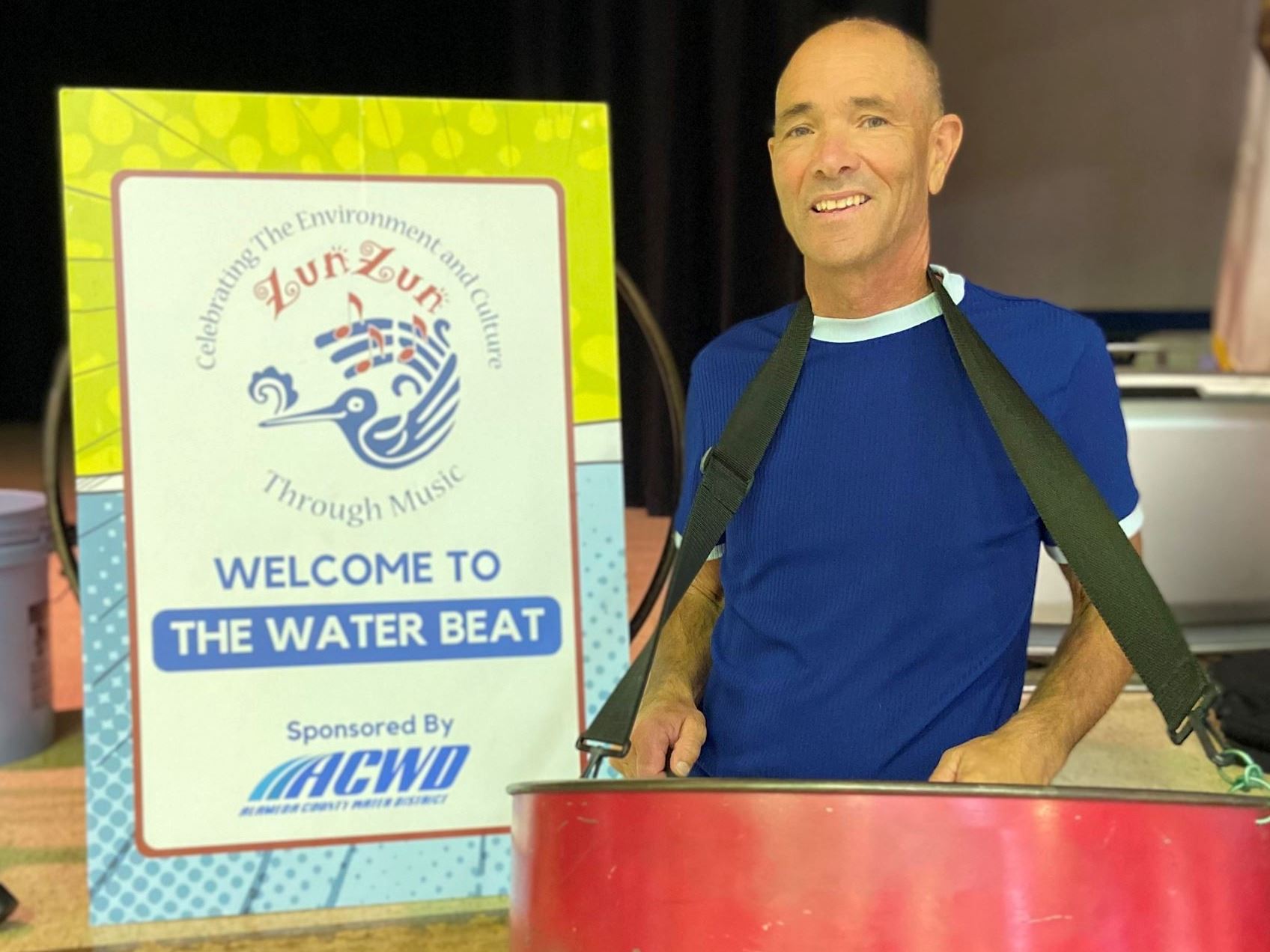 Performer ZunZun kneeling next to musical instruments alongside an a-frame sign with the ZunZun logo